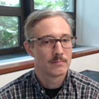 A headshot of a person wearing glasses and a plaid shirt, sitting indoors near a window with greenery visible outside.
