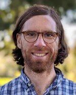 Headshot of a person with glasses, medium-length hair, and a beard, wearing a blue plaid shirt, smiling outdoors in a natural setting.